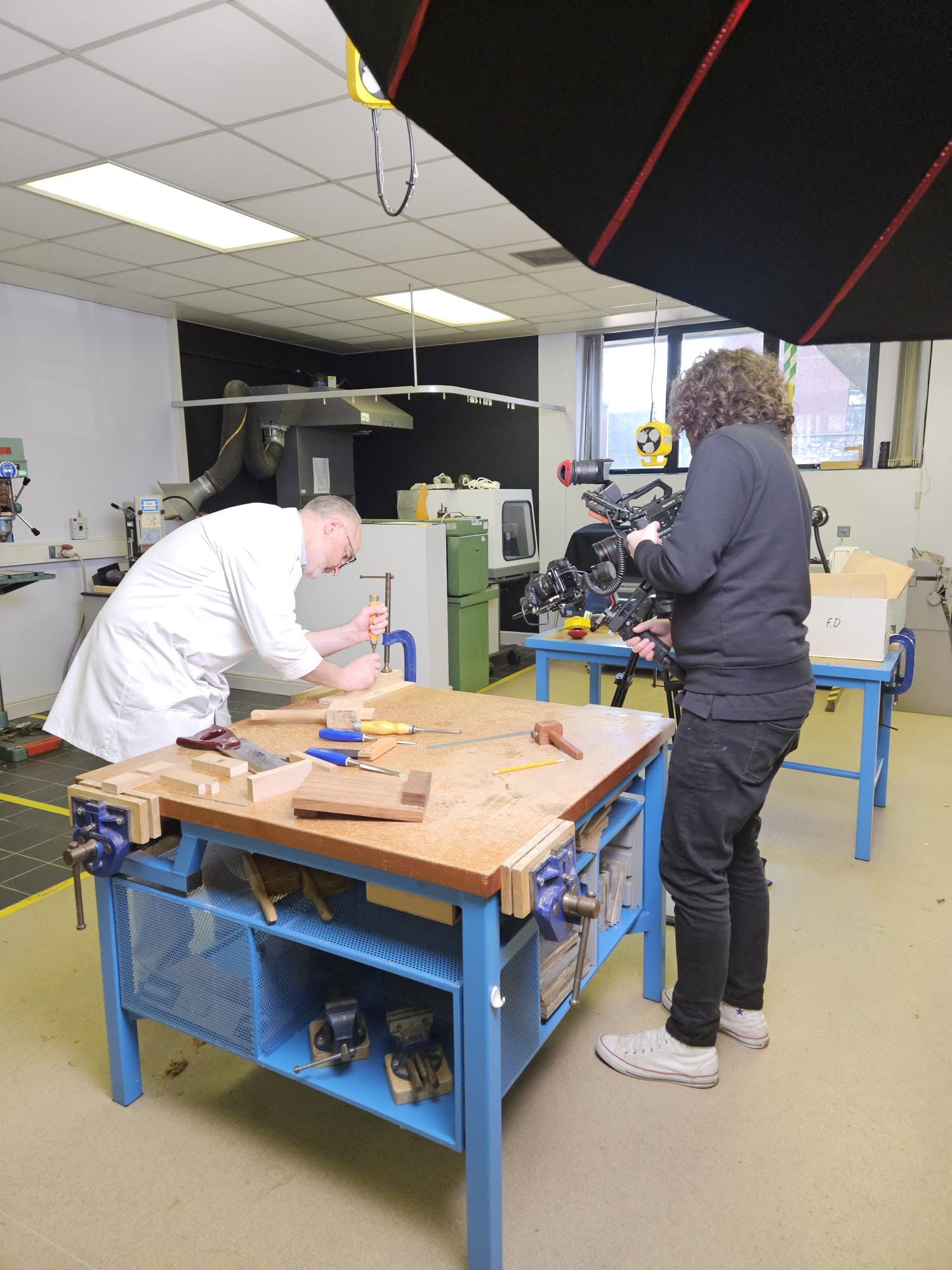 A camera operator films a practical demonstration at a workshop bench.