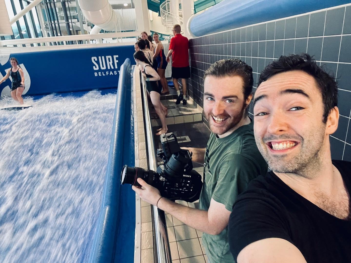 Two members of the SlackPress crew with filming equipment beside the indoor surf pool at Surf Belfast.
