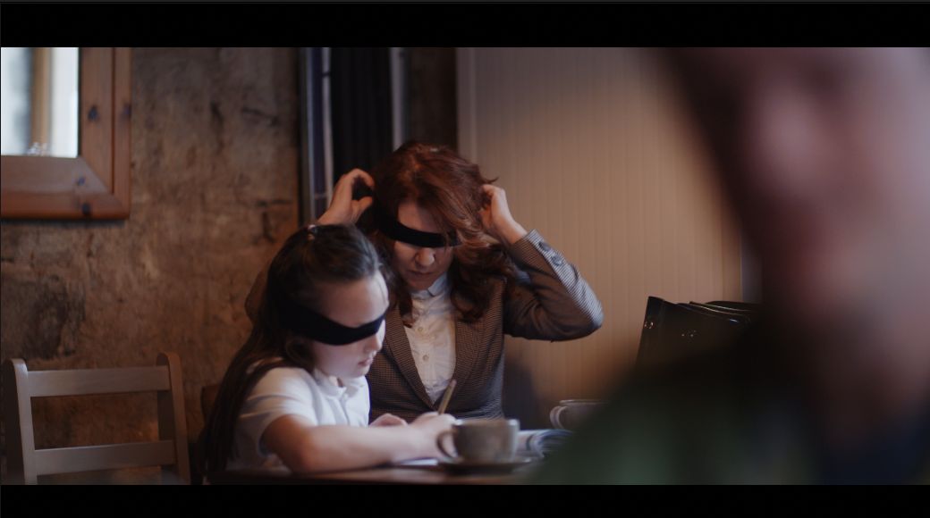 A staged scene from Speak Up showing a blindfolded woman and girl seated at a table.